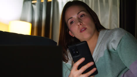 Close-Up-Of-Woman-Sitting-On-Sofa-At-Home-At-Night-Streaming-Or-Watching-Movie-Or-Show-On-Laptop-And-Scrolling-Internet-On-Phone